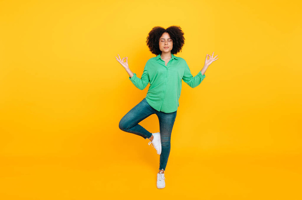 Full length photo of a stylish lovely calm smiling african american or brazilian curly woman meditating while standing tree pose with closed eyes on isolated yellow background. Mental health, harmony - Foto, imagen