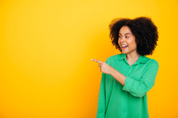 Positive african american or brazilian lovely curly haired woman, wearing green shirt, looks and points with finger to the side at empty space for advertise, smile, stand on isolated yellow background - Photo, Image
