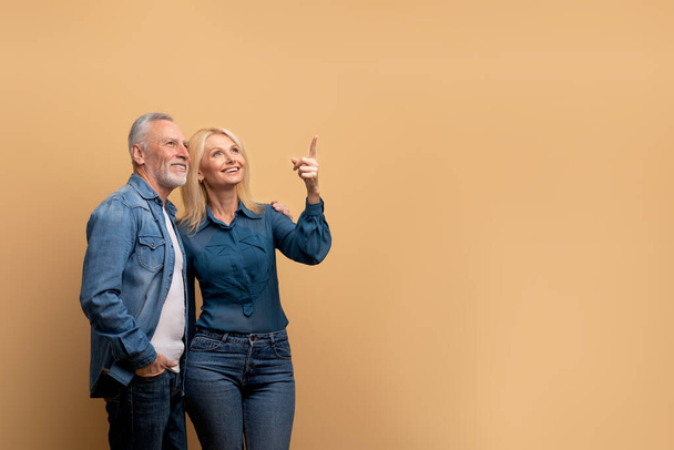 Beautiful cheerful blonde senior woman showing her excited handsome grey-haired bearded husband nice offer, pointing at copy space, old couple isolated on beige studio background - Photo, Image