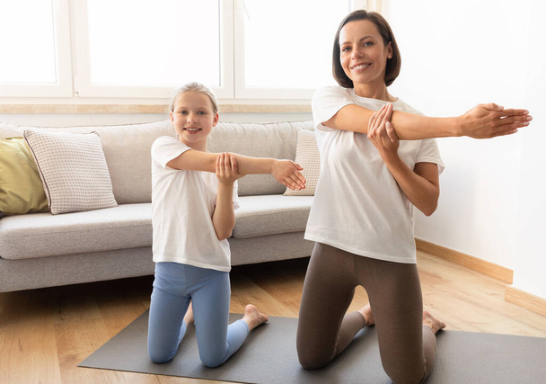 Glückliche kaukasische Frau in Sportbekleidung und Teenager-Tochter praktizieren Yoga und machen Hand-Stretching-Workout im Wohnzimmer Innenraum. Aktiver Lebensstil, Sport zu Hause, gemeinsame Körperpflege - Foto, Bild
