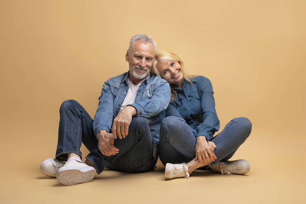 Loving beautiful happy cheerful elderly couple grey-haired man and blonde woman sitting on floor, beige studio background, cuddling, smiling at camera, wearing casual denim outfit. Family portrait - Fotografie, Obrázek