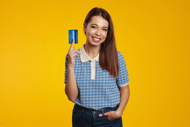 Sonriente chica caucásica muestra tarjeta de crédito, recomienda servicio bancario, se para sobre fondo amarillo, riendo y vistiendo camisa azul - Foto, imagen