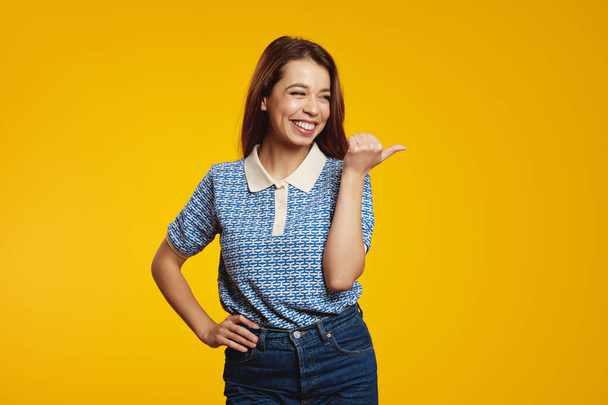 Fröhlich lächelnde Frau zeigt mit dem Finger auf Kopierraum und schaut lächelnd vor gelbem Hintergrund weg. Werbekonzept. - Foto, Bild