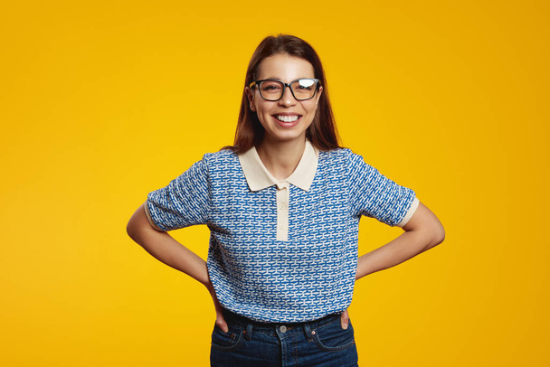 Portrait of cute woman in blue shirt and eyeglasses smiling broadly while holding hands on waist against yellow background - Photo, Image
