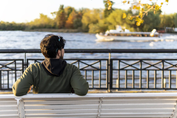 Вид сзади человека, сидящего на скамейке на закате и смотрящего на реку - Фото, изображение