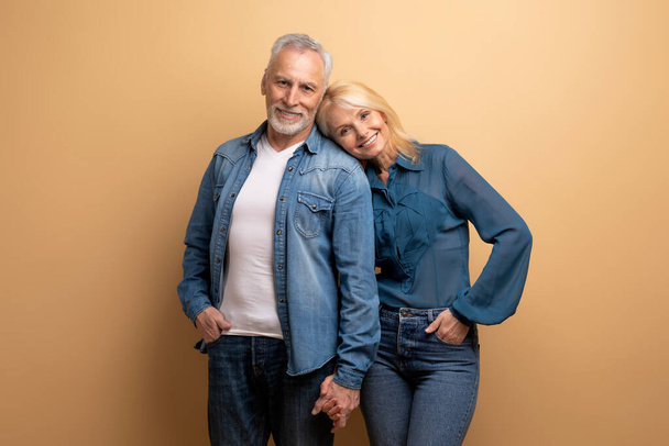 Beautiful loving elderly handsome grey-haired bearded man and pretty blonde woman embracing, holding hands on beige background, smiling at camera. Love, affection, family, marriage - Photo, Image