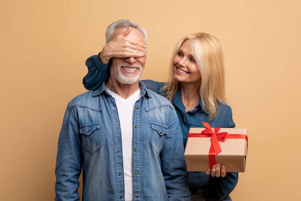 Beautiful cheerful blonde loving mature lady making birthday surprise for husband, senior woman with gift box in her hand covering smiling man eyes, celebrating anniversary together, beige background - Photo, image