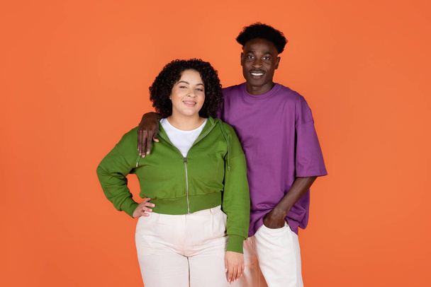 Cool cheerful young mixed race couple african american guy and hispanic curly woman plus size posing together on orange studio background, embracing and smiling at camera - Photo, Image