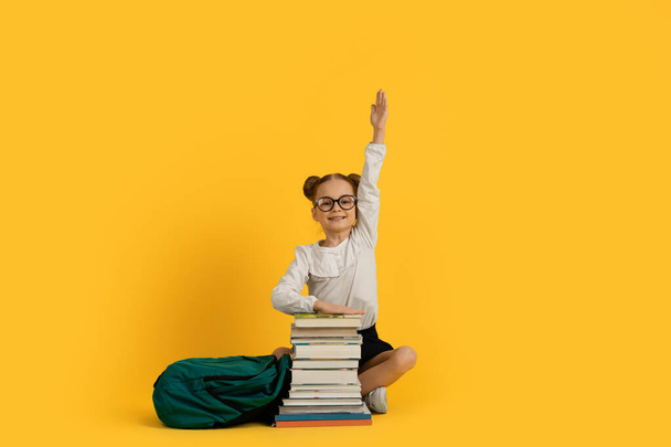Schattig klein Nerdy Schoolmeisje zitten met stapel boeken en raising hand, glimlachen Preteen vrouwelijke kind beantwoorden vraag over les, Genieten van kennis en studie, poseren op gele achtergrond - Foto, afbeelding