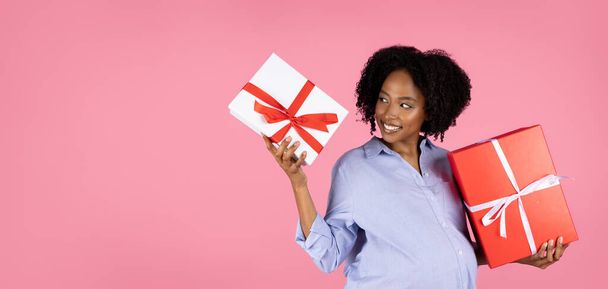 Smiling millennial afrikanisch lockige schwangere Dame mit dickem Bauch hält Schachteln mit Geschenken, isoliert auf rosa Studiohintergrund, Panorama. Wählen Sie Geschenk für den Urlaub, Party zu erwarten Baby - Foto, Bild