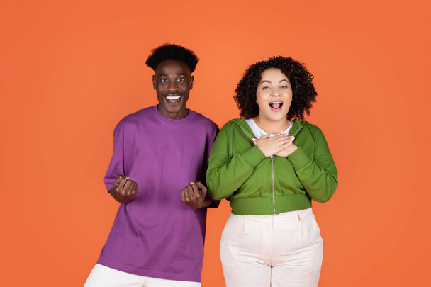 Erstaunlich aufgeregt glücklich junge multikulturelle Paar schöner schwarzer Kerl und hübsche lockige Frau plus Größe gestikuliert und Grimassen auf orangefarbenem Hintergrund, zeigt Glück, Aufregung - Foto, Bild