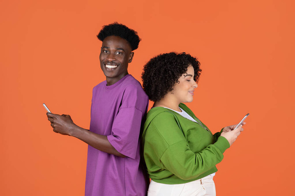 Communication, social media concept. Cheerful interracial young couple in colorful casual outfits standing back to back, using phones and smiling, orange studio background - Photo, Image