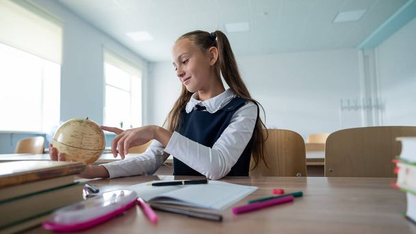 Caucasian schoolgirl sits at her desk at school and studies the globe - Foto, afbeelding