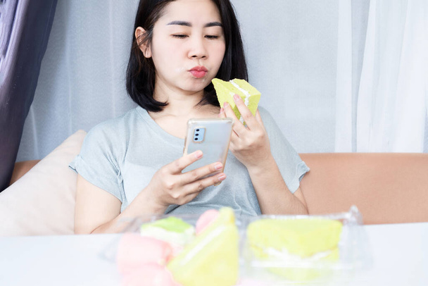 Азиатская женщина, зависимая от сахара и сладостей, ест десерты и пользуется мобильным телефоном - Фото, изображение