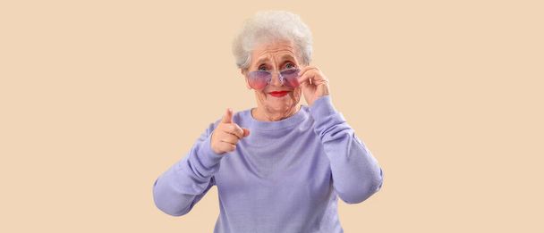 Stilvolle Seniorin mit Sonnenbrille zeigt auf Betrachter auf beigem Hintergrund - Foto, Bild