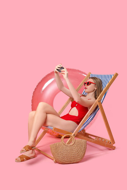 Νεαρή γυναίκα με αξεσουάρ παραλίας που παίρνει selfie σε ξαπλώστρα σε ροζ φόντο - Φωτογραφία, εικόνα