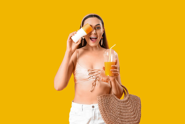Mujer joven con crema protector solar, jugo y bolsa de playa sobre fondo amarillo - Foto, imagen