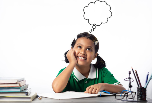 Una bella bambina indiana che studia al tavolo di studio con il viso sorridente che guarda verso l'alto i sogni su sfondo bianco - Foto, immagini