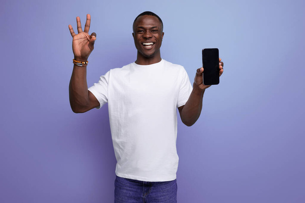 чарівний молодий африканський чоловік у білій футболці тримає смартфон з маком на студійному фоні з копією простору. - Фото, зображення