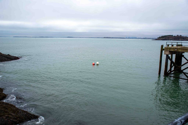 A red and a white buoy float together under an overcast sky along the New England Massachusetts coast line. Boston is visible in the distance. A small dock to the right and a rocky outcropping jetty to the left, the buoys face the open sea. No people - Photo, Image