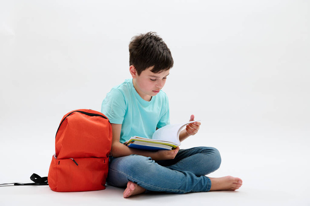 Умный подросток, 11-13 лет, листает в блокноте, делает домашнюю работу, сидит босиком в позе лотоса рядом со своим рюкзаком, изолированный белый студийный фон. Образование. Возвращение в школу - Фото, изображение