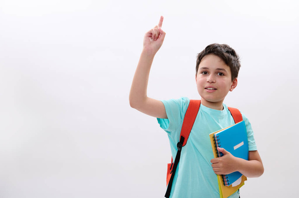 Smart schoolboy, handsome preteen boy with backpack and school supplies, expressing positive emotions, smiles looking at camera, pointing aside a copy space for your ads on white isolated background - Photo, Image