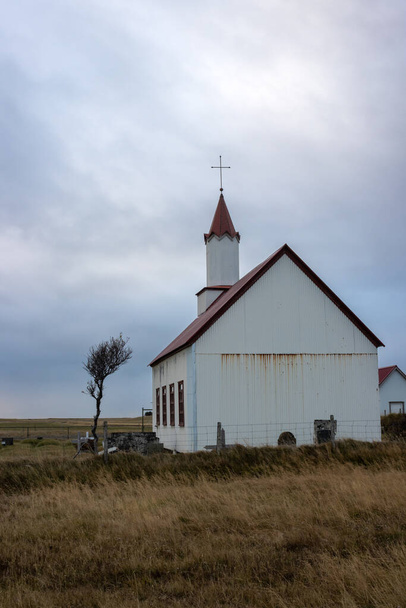 Small white church with a red roof, traditional icelandic combination. Small tree beside. Intense cloudy sky. Autumn color of the grass. South Iceland. - Foto, Imagen