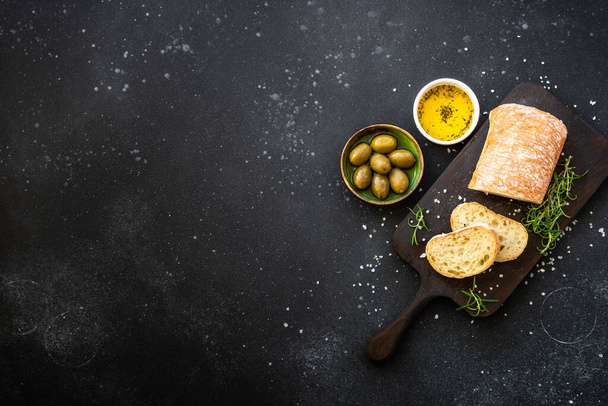 Pão Ciabatta sobre tábua de madeira com azeite, azeitonas e ervas em preto. Vista superior com espaço de cópia. - Foto, Imagem