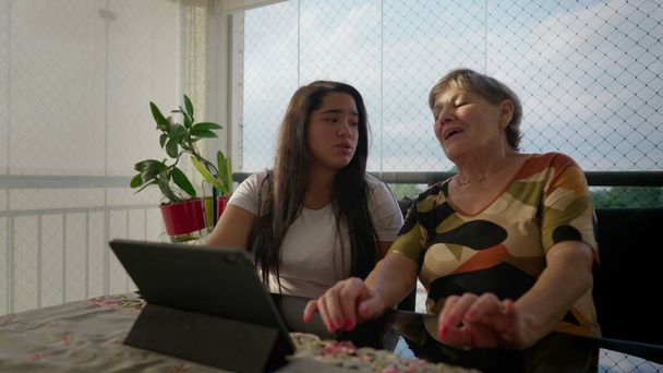 Alegre abuela en frente del dispositivo de tableta con su nieta asiática diversa en balcón apartamento. Escena intergeneracional de auténtico momento familiar diverso - Foto, imagen
