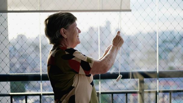 Счастливая пожилая женщина стоит у балкона квартиры, открывая жалюзи, чтобы насладиться дневным солнцем. Старший пенсионер закрывает жалюзи - Фото, изображение