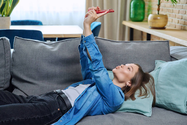 Young woman having video call chat conference lying on sofa at home using smartphone. Female recording video blog making online broadcast talking with friends study work lifestyle mobile applications - 写真・画像
