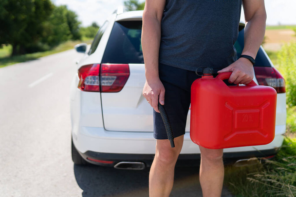 Een man houdt een rood blik vast naast een witte auto. Stop om bij te tanken, problemen op de weg met tanken - Foto, afbeelding