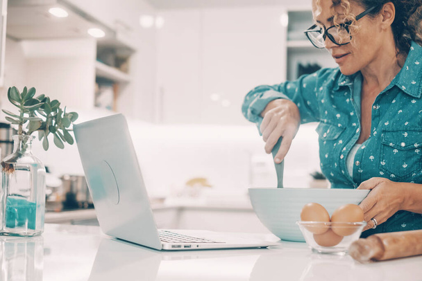 Seitenansicht der erwachsenen Frau glücklich lächelnd und Vorbereitung hausgemachten Kuchen mit Eiern und Mehl in handgemachten klassischen Stil folgenden Anweisungen auf Laptop-Computer mit Online-Tutorial-Klasse.  - Foto, Bild