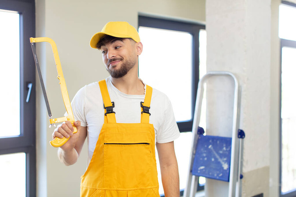 young handsome man smiling and looking with a happy confident expression. handyman with a saw - Фото, изображение