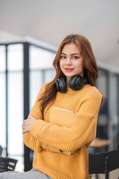 Portret atrakcyjnej młodej Azjatki w przytulnym żółtym swetrze stoi w kawiarni z książką i słuchawkami. studentka, freelancer.. - Zdjęcie, obraz