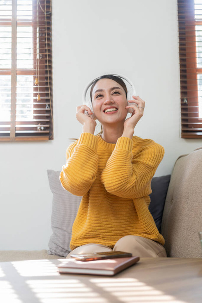 Young beautiful Asian girl in yellow shirt, happy carefree wearing headphones enjoying relaxing music in living room with weekend at home. - Zdjęcie, obraz