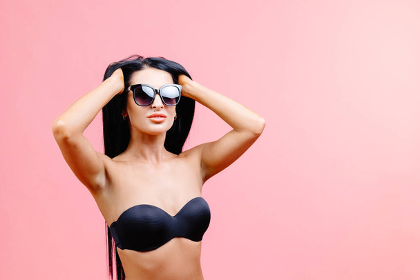 Junge Frau im sommerlichen schwarzen Badeanzug und Sonnenbrille auf rosa Hintergrund im Studio. - Foto, Bild