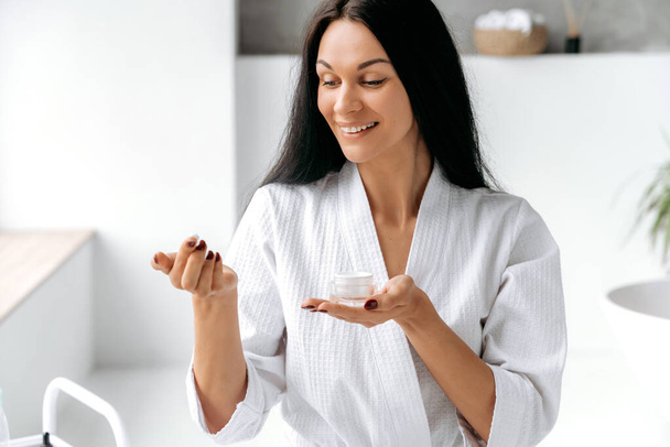 Beautiful well-groomed caucasian long-haired woman in a white bathrobe, sits at home in the bathroom, holds a moisturizing face cream in her hand, prepares for a skin care procedure, smile - Foto, Imagen