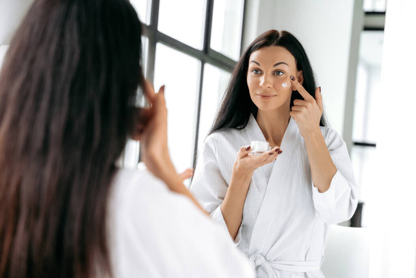 Upea valkoihoinen brunette pitkäkarvainen nainen valkoisessa kylpytakissa seisoo kylpyhuoneessa peilin edessä, soveltaen kosteusvoidetta kasvoihin, hymyillen. Ihon ravitsemus ja nesteytys - Valokuva, kuva