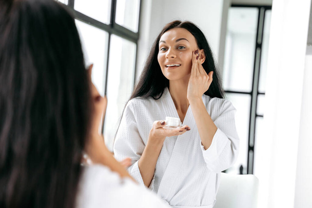 Beauty-Konzept. Wunderschöne kaukasische brünette langhaarige Frau in einem weißen Bademantel steht im Badezimmer vor einem Spiegel, trägt Feuchtigkeitscreme ins Gesicht, lächelt. Ernährung und Hydratation der Haut - Foto, Bild