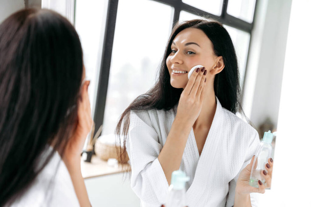 Skin cleansing. Lovely caucasian brunette woman, in a bathrobe, stands in the bathroom in front of a mirror, cleans her face after makeup using cotton pads and tonic, smile - Photo, Image