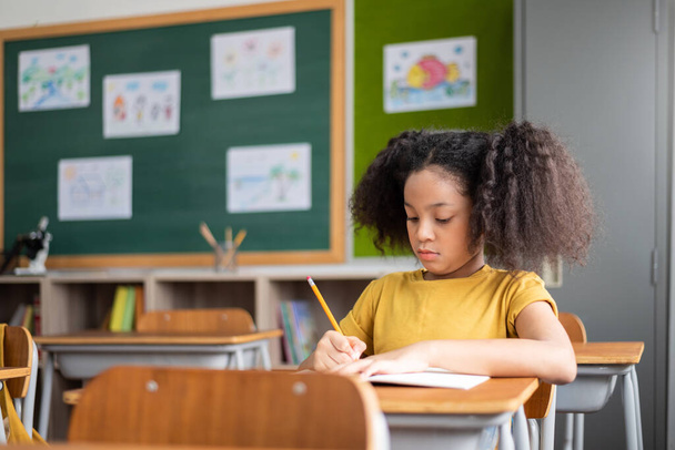 Concentrated African American girl with pigtails doing exam at classroom. Child sitting at desk writing in notebook. Education, Elementary school. - Foto, Bild