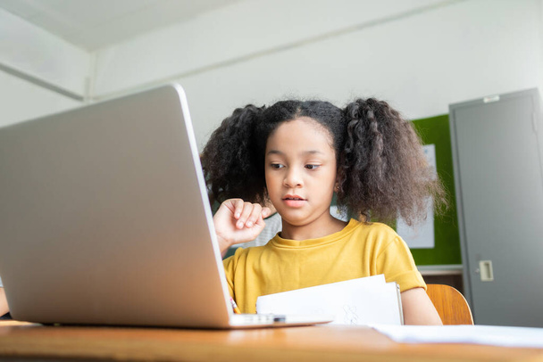 African American Elementary schoolgirl with pigtails studying with laptop computer. Browsing Internet, Modern classroom, Education, Online Learning, E-Learning Concept - Фото, изображение