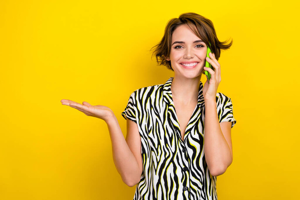 Photo of optimistic pleasant lovely girl palm demonstrate offer empty space talking on smartphone isolated on yellow color background. - Foto, immagini