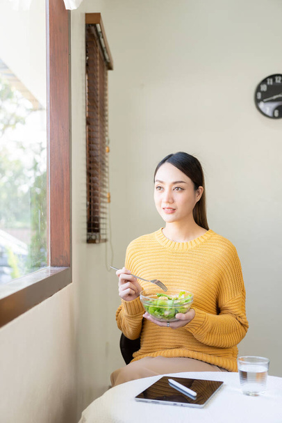 Счастливая молодая азиатская женщина со свежими овощами и фруктами, здоровый рабочий обед дома, красивая женщина с салатом для ноутбука, образ жизни в гостиной. - Фото, изображение