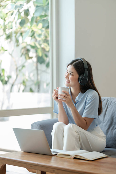 Young woman relaxing and drinking cup of hot coffee or tea using laptop computer on sofa at living room. High quality photo - Foto, afbeelding