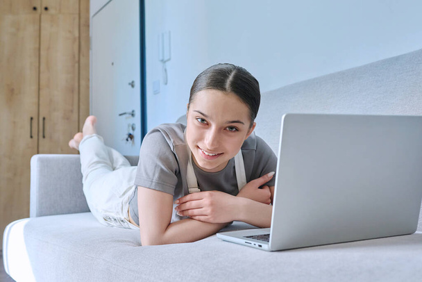 Teen girl at home lying on couch using laptop, smiling looking at camera. Technology for learning leisure communication, modern lifestyle, adolescence concept - Valokuva, kuva