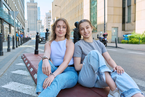 İki gülümseyen genç kız öğrenci kameraya bakıyor, modern şehir tarzı, binaların arka planı. Moda kızları yol kenarında kaldırımda oturuyorlar. Gençlik, moda, tarz, şehir, yaşam tarzı konsepti - Fotoğraf, Görsel