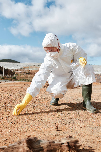 Environmental researcher in mask and safety goggles taking sample of soil and putting it into lastic bag on polluted land - 写真・画像