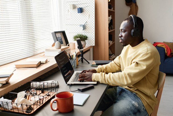 Concentrated young African-American coding specialist in headphones sitting at table against window and using laptop while creating app - Valokuva, kuva